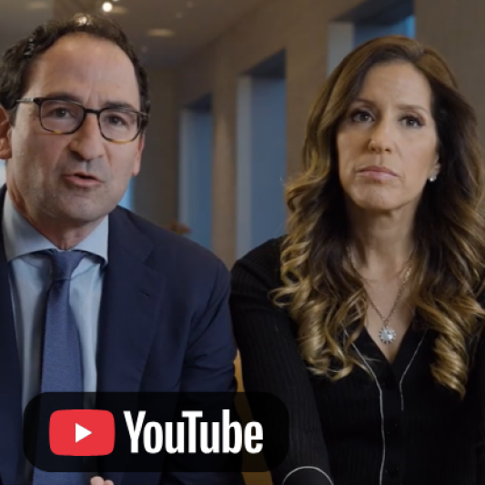 Video thumbnail featuring two people seated indoors and speaking to the camera, with a YouTube icon in the lower-left corner.