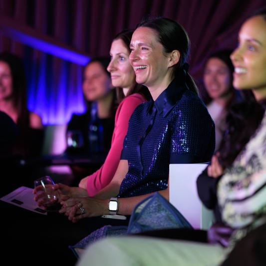 Audience members seated at an indoor event, watching a presentation under colorful stage lighting.